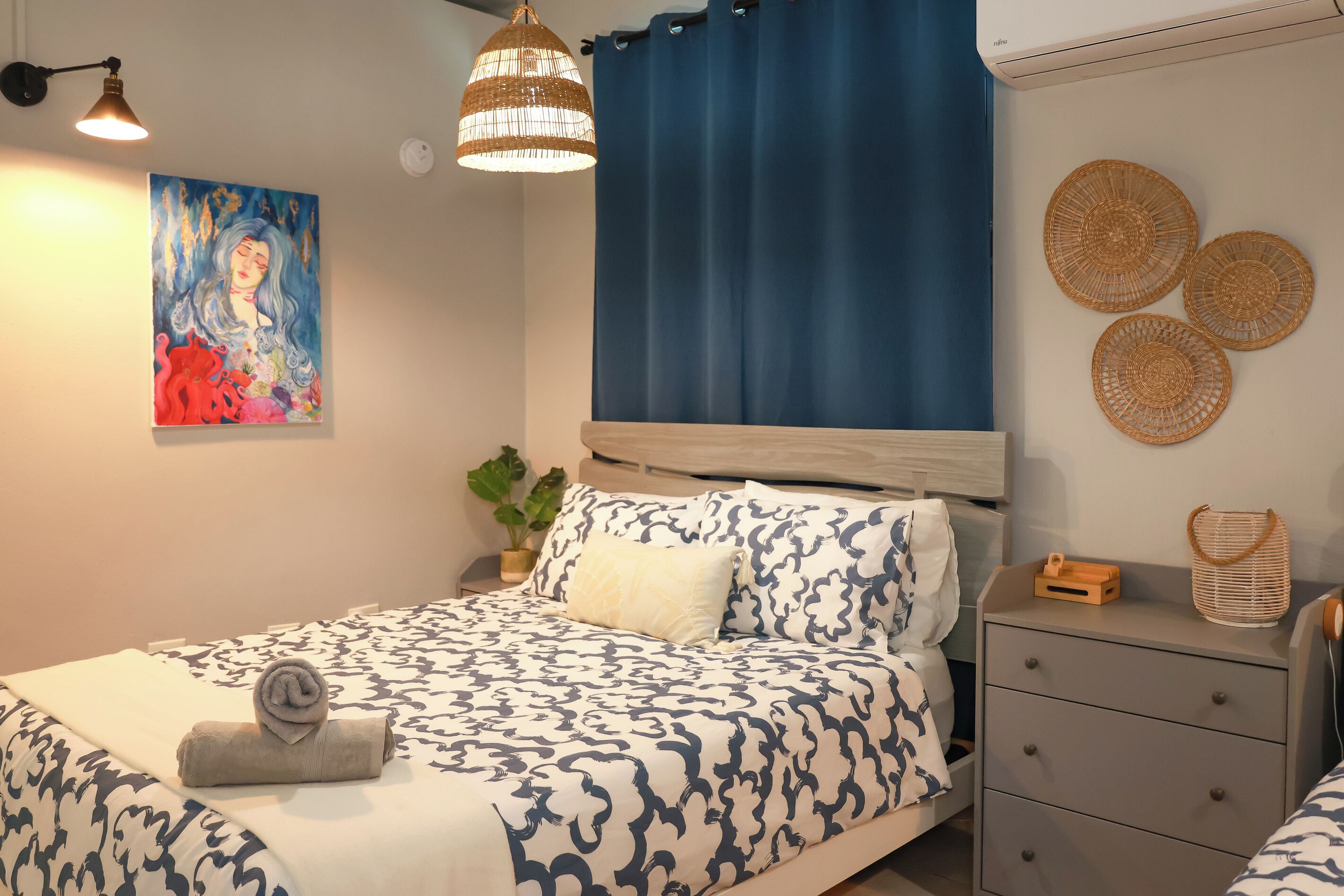 A bedroom with a bed, bedside table, lamp, artwork, and decorative wall hangings.