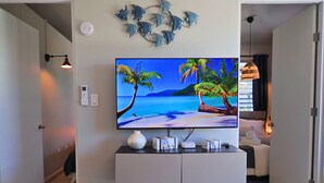 A modern living room with a flat-screen TV mounted on a white cabinet, a decorative wall art of fish, and a view of a beach scene on the TV screen. - Casa Arrecife Queen Beds/Beach/Pool/Pet Friendly (Cabo Rojo)