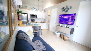 A modern living room with a blue sofa, a TV mounted on the wall, and a kitchen area with white cabinets and a microwave. - Casa Arrecife Queen Beds/Beach/Pool/Pet Friendly (Cabo Rojo)