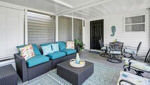 Een overdekte veranda met een donkere rieten bank, turquoise kussens en een kleine tafel met een vaas.