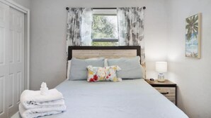 Een slaapkamer met een bed, een nachtkastje, een raam met gordijnen, een deur en een aan de muur hangend schilderij.