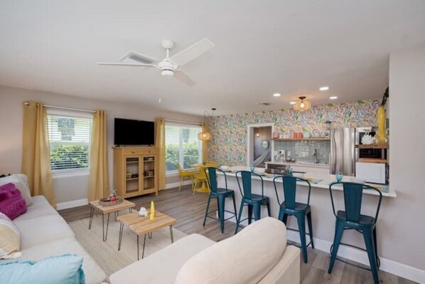 Un salon moderne avec un canapé, une table basse et des fauteuils, une cuisine avec un coin repas et un ventilateur de plafond.
