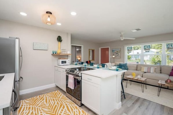 A modern kitchen with white cabinets, a built-in oven, and a microwave. There is a dining area with a sofa and a coffee table.
