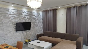 Ein Wohnzimmer mit braunem Sofa, weißem Couchtisch, einem flachbildfernseher an einer strukturierten Wand und einem Esstisch mit Tischdecke mit Obstmuster.