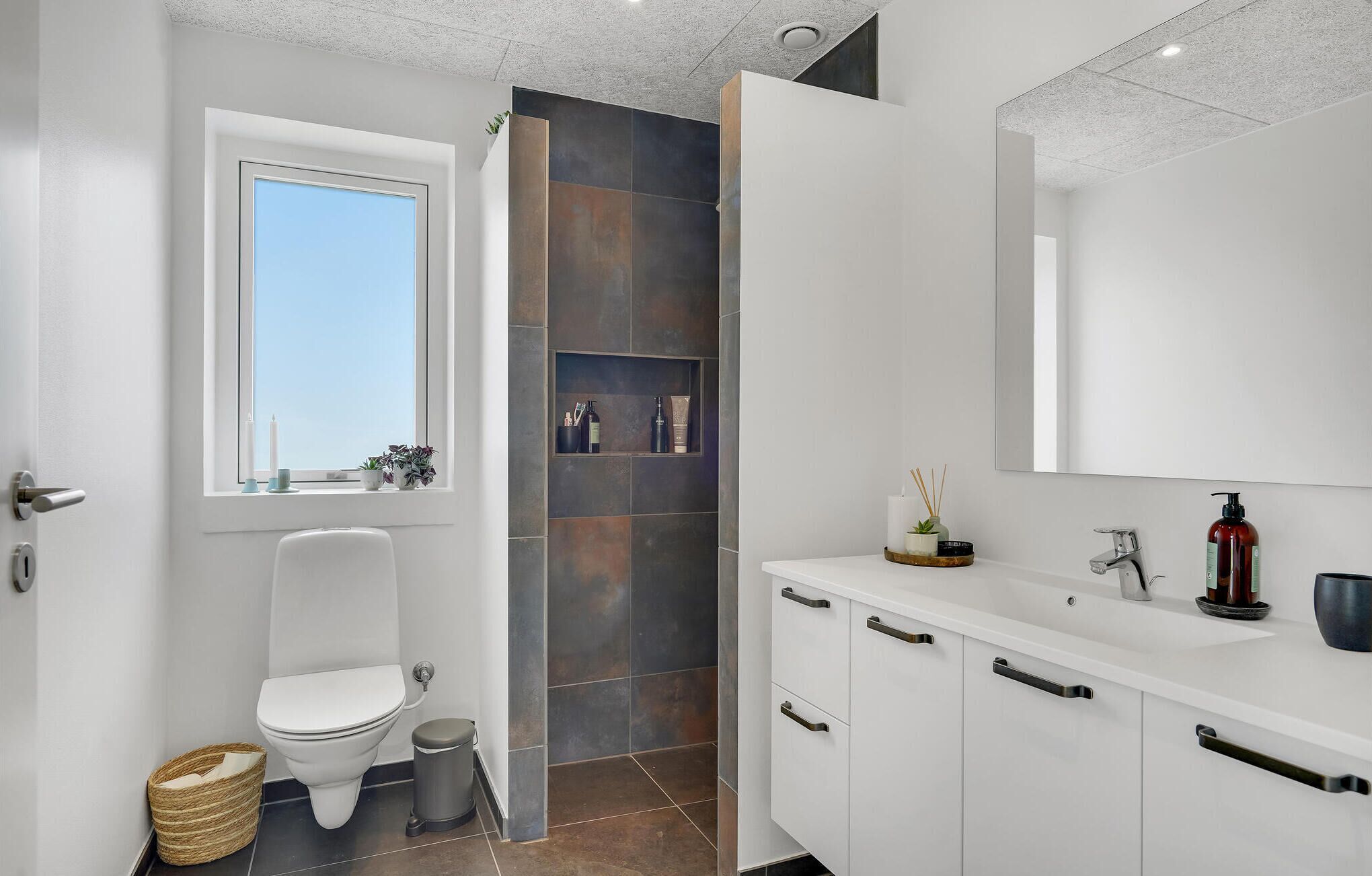 A modern bathroom with a toilet, sink, and mirror. There is a window above the toilet and a shelf with toiletries.