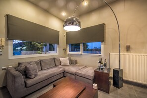 Un salon moderne avec un canapé d’angle, une table basse en bois, une table d’appoint en verre sur laquelle sont posées des bouteilles, et un grand luminaire suspendu.
