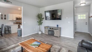 Ein modernes Wohnzimmer mit einem Holztisch, einem flachbildfernseher an der Wand und einer Küchenzeile im Hintergrund.