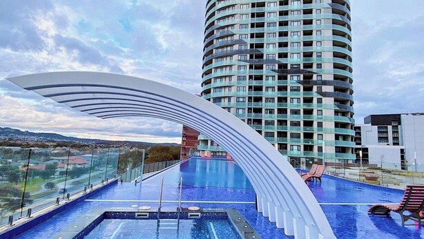 A modern hotel with a curved swimming pool and a view of a tall building in the background. - Spacious 1 Bed Apartment, parking, gym, pool (Gungahlin)