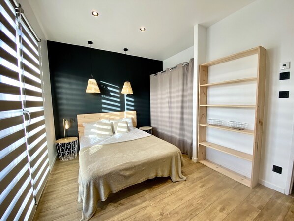 Quarto moderno com piso de madeira, cama com cobertura bege, duas luminárias pendentes, parede escura e um conjunto de prateleiras de madeira.