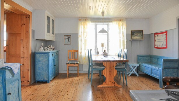 A dining area with a wooden table, chairs, and a blue bench. There is a window with curtains, a door, and a wall with a framed picture.