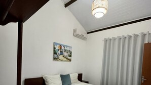 A bedroom with a bed, a nightstand, a painting, an air conditioning unit, and a window with curtains. - Suítes D'Ajuda Inn (Porto Seguro)
