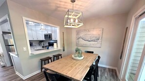 Un coin repas avec une table en bois, des chaises noires, un lustre, une cuisine en arrière-plan et un grand tableau au mur.