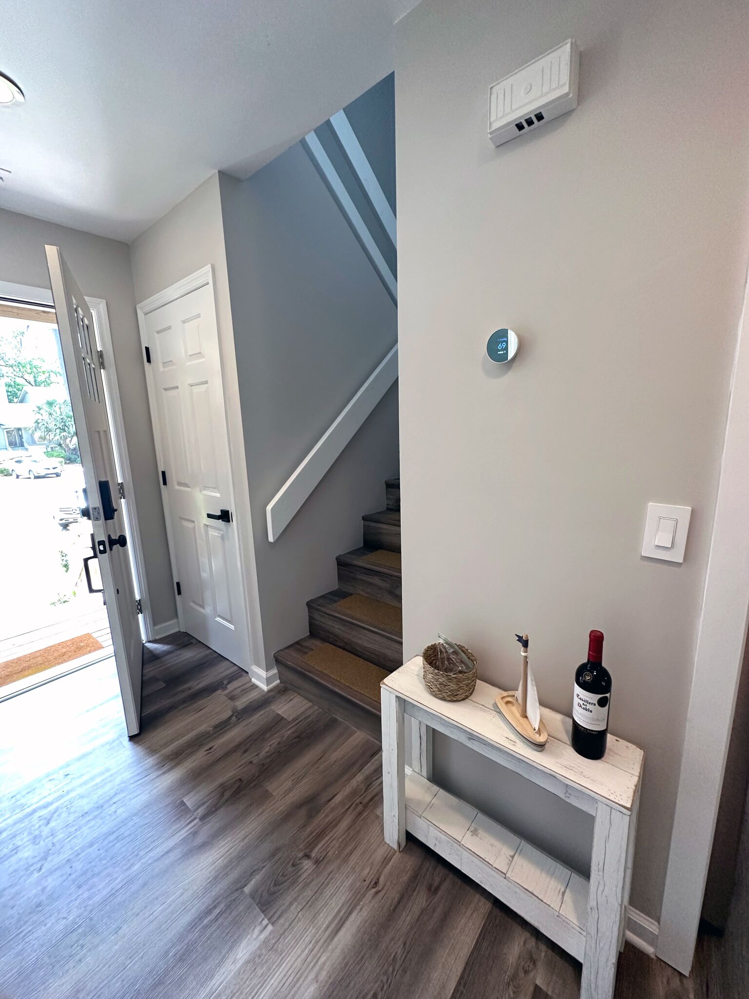 A hallway with a staircase, a white door, a wall-mounted thermostat, and a small table with a wine bottle and decorative items.