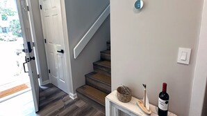 Un couloir avec un escalier, une porte blanche, un thermostat fixé au mur et une petite table sur laquelle se trouvent une bouteille de vin et des objets décoratifs.