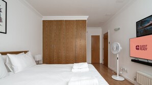 A hotel room with a large bed, wooden wardrobe, a TV displaying "GUEST READY", and a standing fan. - GuestReady - Trendy stay near Brick Lane Market (London)