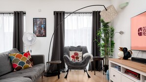 A modern living room with a grey sofa, a black floor lamp, a grey armchair with a decorative pillow, a white side table, a wooden TV stand, and a potted plant. - GuestReady - Trendy stay near Brick Lane Market (London)