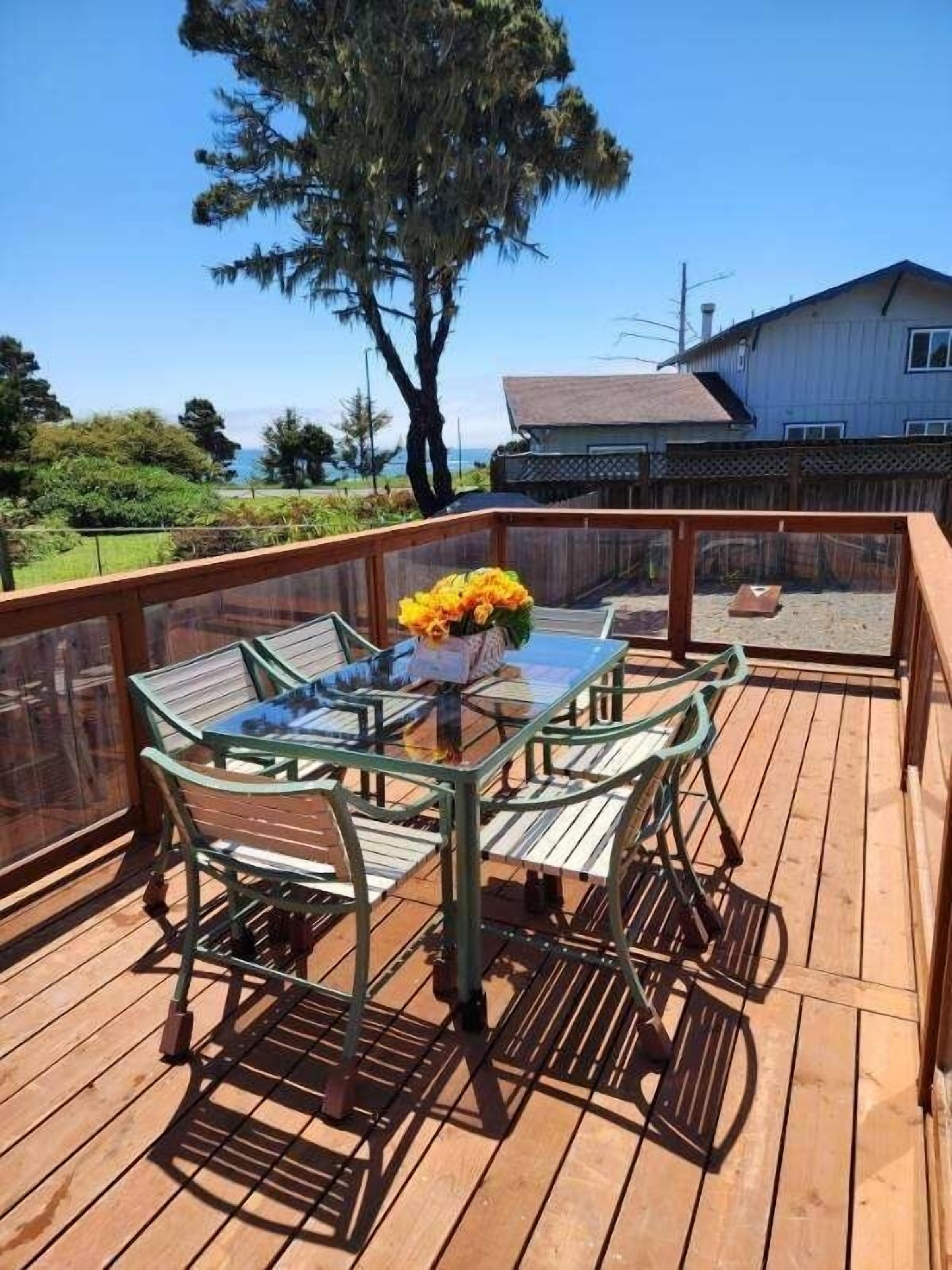 A wooden deck with a glass table and chairs, a flower arrangement, and a view of a house and trees.