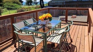 Une terrasse en bois avec une table et des chaises en verre, un arrangement floral et une vue sur une maison et des arbres.