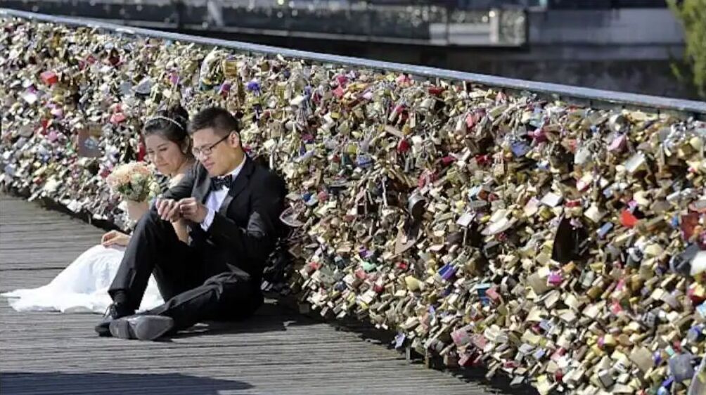 Un couple de jeunes mariés est assis sur une terrasse en bois, avec un mur couvert de cadenas derrière eux.