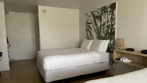 A hotel room with two beds, a wooden floor, a wall-mounted shelf with a fan, and a door leading to another room.