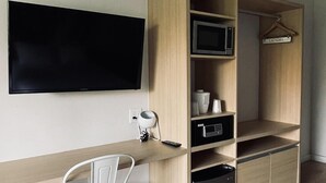 A compact room with a wooden desk, a white chair, a television mounted on the wall, and a built-in wardrobe with a hanging space.