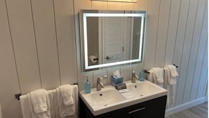 A bathroom with a vanity, two sinks, a mirror with lights, and towels on the wall.