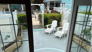 A view through a window of a patio area with white chairs, a wooden deck, and a white fence.