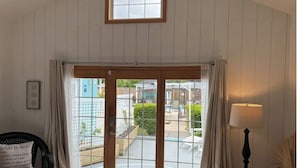 A room with a large sliding glass door leading to a balcony with a view of a residential area. There is a chair with a cushion, a lamp, and a framed picture on the wall.