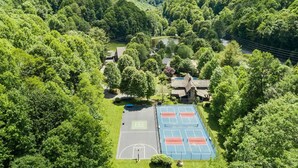 An aerial view of a property surrounded by lush green hills, featuring a tennis court and a building with a sloped roof. - Sunset Mtn View*Pool*Hot Tub*Massage Chair*Theater Rm*Pickleball*Game Rm*Hiking (Mars Hill)