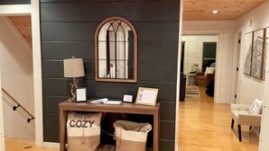 A hallway with a dark wall, a wooden console table with a lamp, framed certificates, and two wicker baskets labeled "COZY". - Sunset Mtn View*Pool*Hot Tub*Massage Chair*Theater Rm*Pickleball*Game Rm*Hiking (Mars Hill)