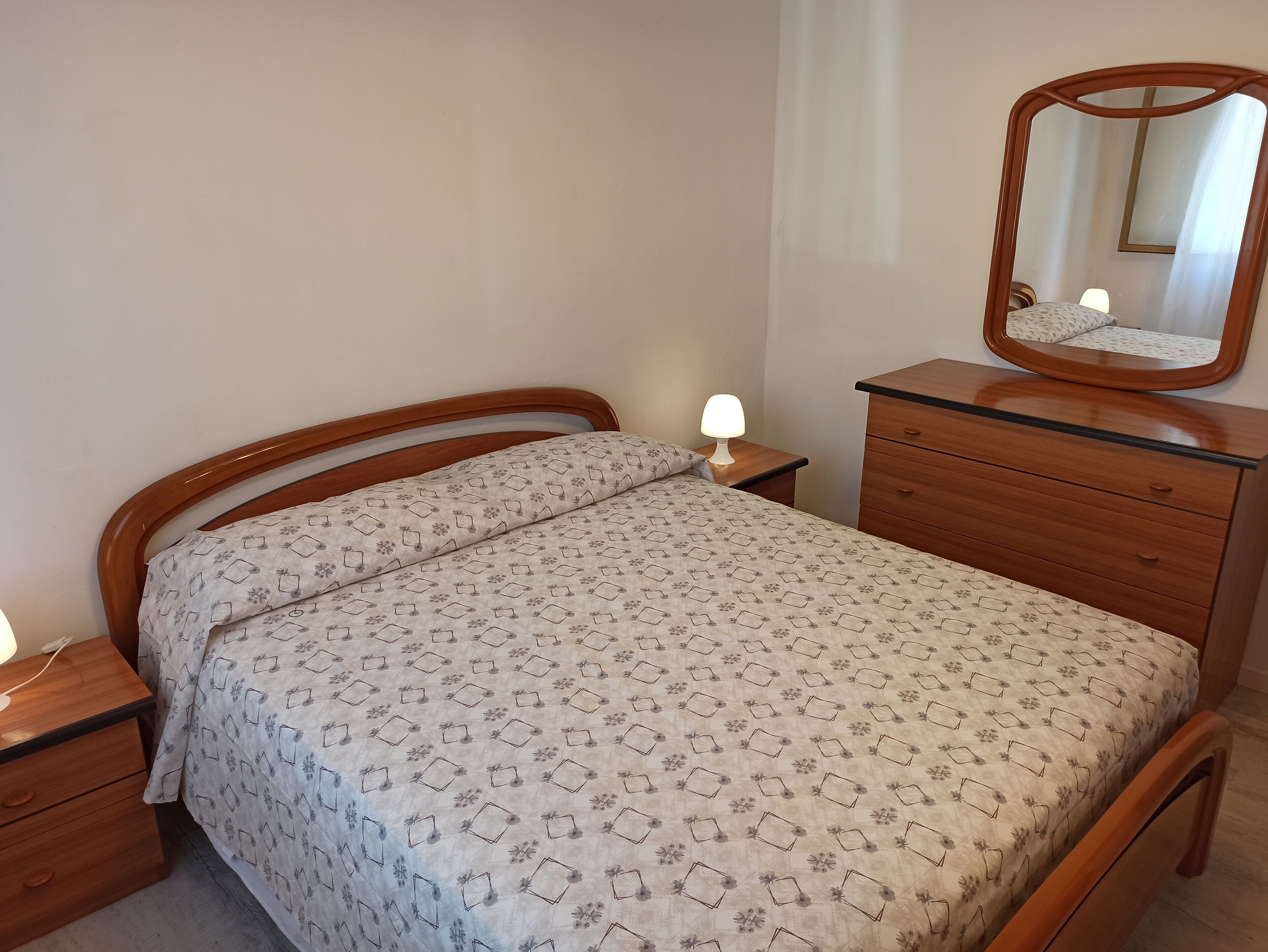 Ein Schlafzimmer mit einem hölzernen Bett, einem Nachttisch mit Lampe, einer Kommode mit Schubladen und einem Spiegel auf der Kommode.