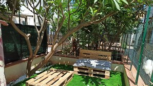 Ein kleiner Sitzbereich im Freien mit einer Holzbank und einem Tisch, umgeben von einem grünen Kunstrasen und einem Baum.