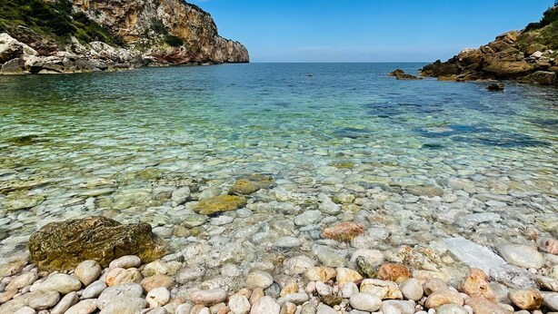 Ein felsiger Strand mit klarem, flachem Wasser und einer gebirgigen Kulisse.