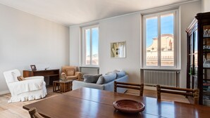 Ein großzügiger Wohnbereich mit Esstisch aus Holz, einer Couch und einem Sessel. Große Fenster bieten Blick auf die Stadtlandschaft.