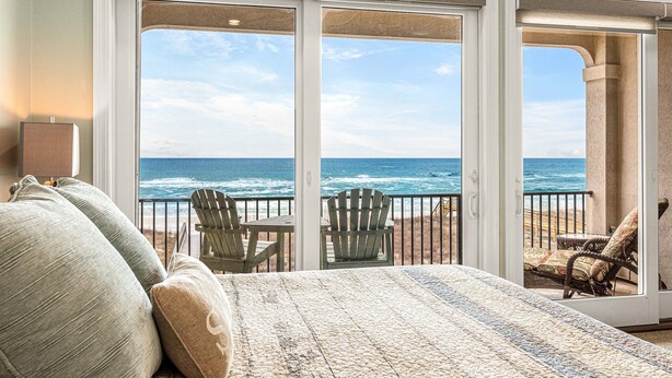 Une chambre avec une grande porte-fenêtre coulissante donnant sur un balcon avec vue sur l’océan. On y trouve deux chaises en bois et une petite table.