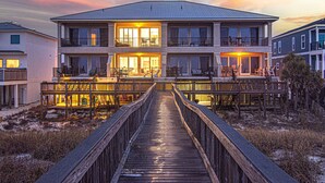 Une maison à deux étages avec une terrasse en bois menant à l’entrée, entourée d’un paysage sablonneux.