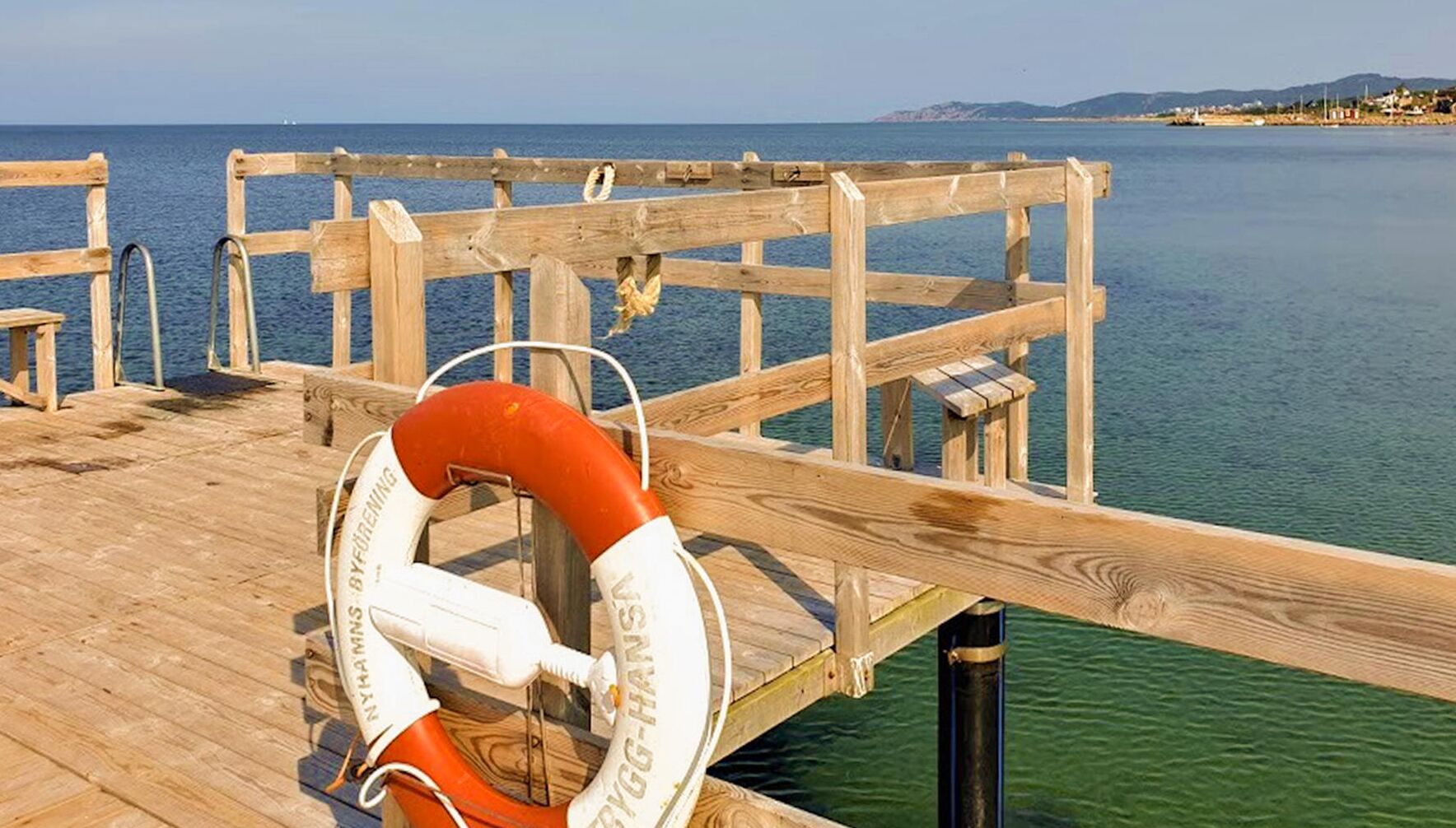 Eine hölzerne Anlegestelle erstreckt sich über ruhiges Wasser, an der Geländer eine Rettungsboje befestigt ist.