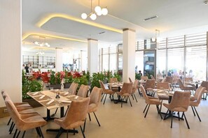 A modern restaurant with round tables and cushioned chairs, large windows, and indoor plants. - FLLAD RESORT & SPA (Golem)