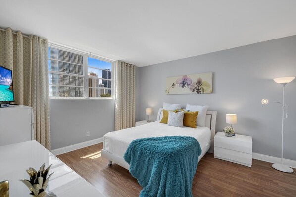 A modern hotel room with a bed, bedside tables, a TV, and a view of the cityscape.