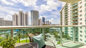 A balcony with a view of high-rise buildings, a palm tree, and a chair with cushions. - Ocean View, 1BD+1.5BA, Steps To Beach. Heated Pool. Ocean reserve (Sunny Isles Beach)