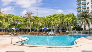 A swimming pool with lounge chairs and umbrellas, surrounded by palm trees and a building with balconies. - Ocean View, 1BD+1.5BA, Steps To Beach. Heated Pool. Ocean reserve (Sunny Isles Beach)