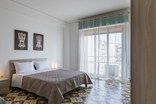 Un dormitorio con una cama, una mesita de noche, una ventana con cortinas traslúcidas y dos obras de arte colgadas en la pared.
