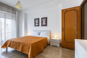 Quarto com cama, mesas de cabeceira, um criado-mudo, porta de madeira e duas obras de arte penduradas na parede.