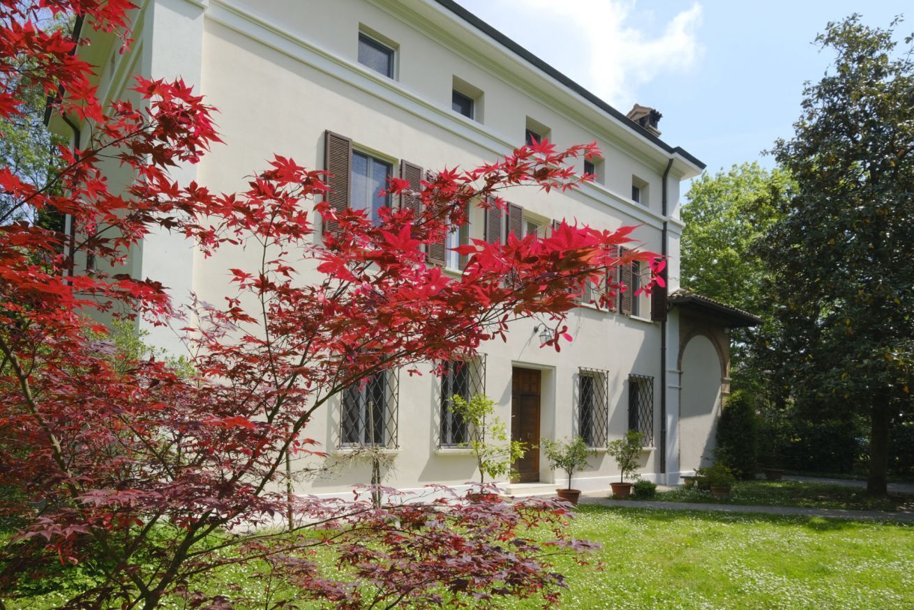 A two-story building with a white facade, multiple windows with shutters, and a small garden area with red foliage.