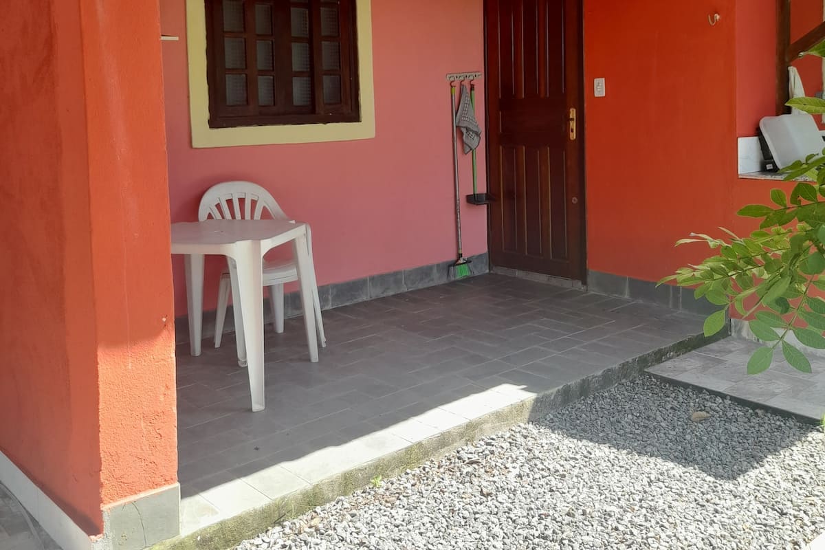 Uma pequena construção térrea com piso de azulejos, porta de madeira, janela e uma cadeira de plástico branca.