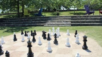 Un grand jeu d’échecs en plein air est installé sur une pelouse, avec des marches menant à un mur de pierre en arrière-plan.