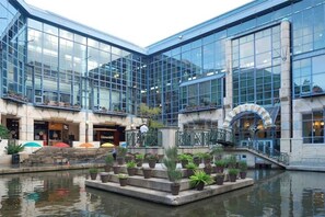 Ein modernes Gebäude mit großen Glasfenstern, einem zentralen Innenhof mit Wasserspiel und einer Treppe, die zu einer oberen Ebene führt.