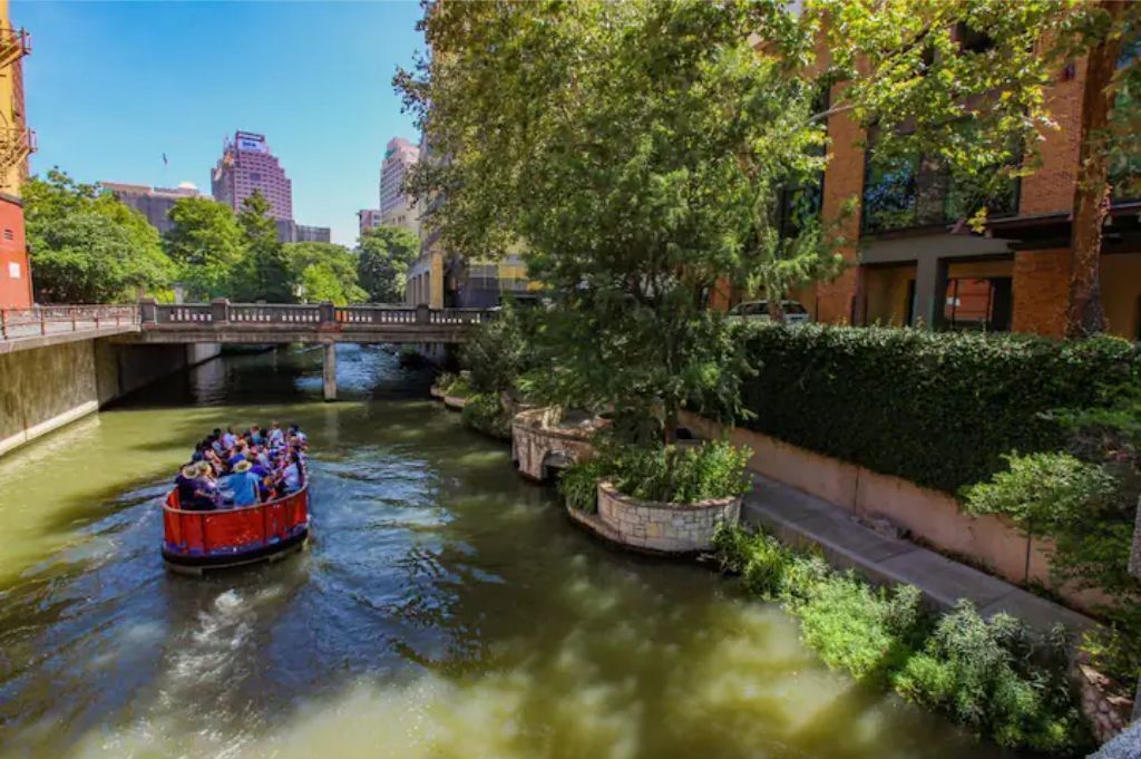 A river with a boat carrying passengers, surrounded by greenery and buildings.