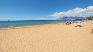 Une vaste plage de sable fin, avec une eau d’un bleu limpide et quelques personnes aperçues au loin.
