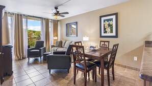 A hotel room with a dining area, a ceiling fan, a large window with curtains, and two framed pictures on the wall.